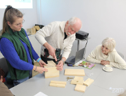 Zdjęcie przedstawia starszego mężczyznę, który wierci w desce wiertarką. Obok niego przy stole siedzi starsza kobieta. Deskę trzyma młoda dziewczyna.