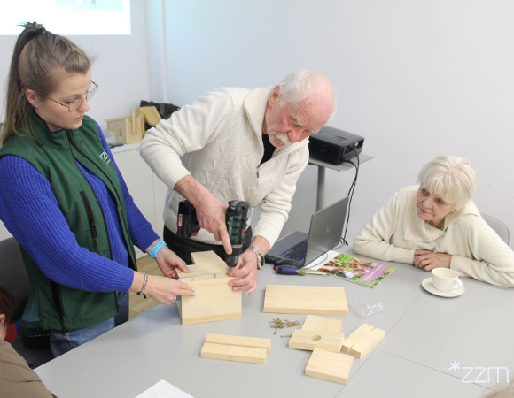 Zdjęcie przedstawia starszego mężczyznę, który wierci w desce wiertarką. Obok niego przy stole siedzi starsza kobieta. Deskę trzyma młoda dziewczyna.