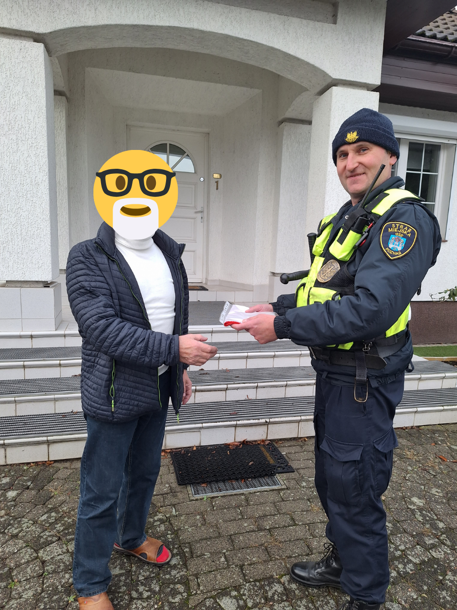 zdjęcie przedstawia strażnika miejskiego przed domem wręczającego apakowanie ze złożoną flagą Polski mężczyźnie w białym swetrze i czarną kurtką, twarz mężczyzny zakrywa emotikonka uśmiechu w okularach i z białą brodą