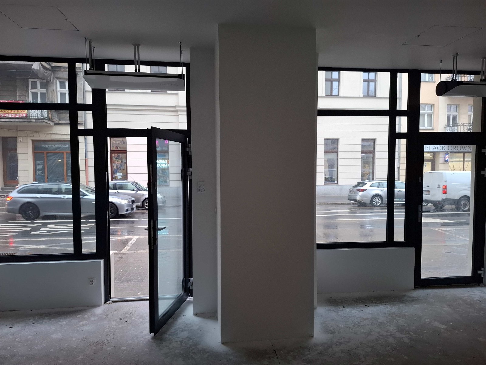Zdjęcie przedstawia surowe wnętrze lokalu użytkowego przeznaczonego na wynajem, w którym dominują duże, przeszklone witryny w czarnych obramowaniach, wychodzące bezpośrednio na ruchliwą ulicę. Przez otwarte drzwi i szyby widoczna jest mokra nawierzchnia jezdni, parkujące samochody oraz elewacje sąsiednich kamienic, w tym szyld salonu "BLACK CROWN".