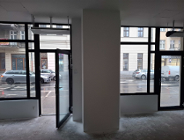 Zdjęcie przedstawia surowe wnętrze lokalu użytkowego przeznaczonego na wynajem, w którym dominują duże, przeszklone witryny w czarnych obramowaniach, wychodzące bezpośrednio na ruchliwą ulicę. Przez otwarte drzwi i szyby widoczna jest mokra nawierzchnia jezdni, parkujące samochody oraz elewacje sąsiednich kamienic, w tym szyld salonu "BLACK CROWN".