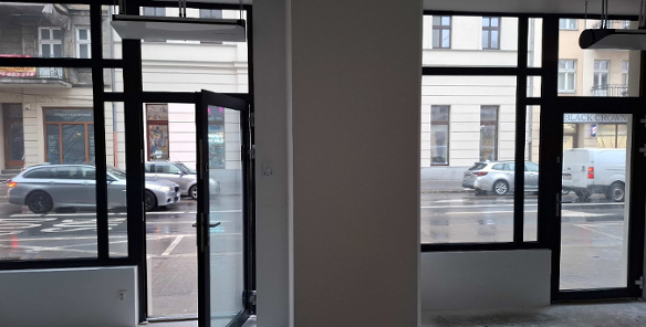 Zdjęcie przedstawia surowe wnętrze lokalu użytkowego przeznaczonego na wynajem, w którym dominują duże, przeszklone witryny w czarnych obramowaniach, wychodzące bezpośrednio na ruchliwą ulicę. Przez otwarte drzwi i szyby widoczna jest mokra nawierzchnia jezdni, parkujące samochody oraz elewacje sąsiednich kamienic, w tym szyld salonu "BLACK CROWN".