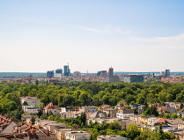 Zdjęcie przedstawia szeroką panoramę Poznania, ukazaną z wysokiego punktu obserwacyjnego, gdzie na pierwszym planie dominują dachy niskiej zabudowy mieszkalnej otoczonej gęstą, soczystą zielenią drzew.