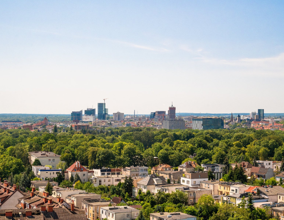 Zdjęcie przedstawia szeroką panoramę Poznania, ukazaną z wysokiego punktu obserwacyjnego, gdzie na pierwszym planie dominują dachy niskiej zabudowy mieszkalnej otoczonej gęstą, soczystą zielenią drzew.