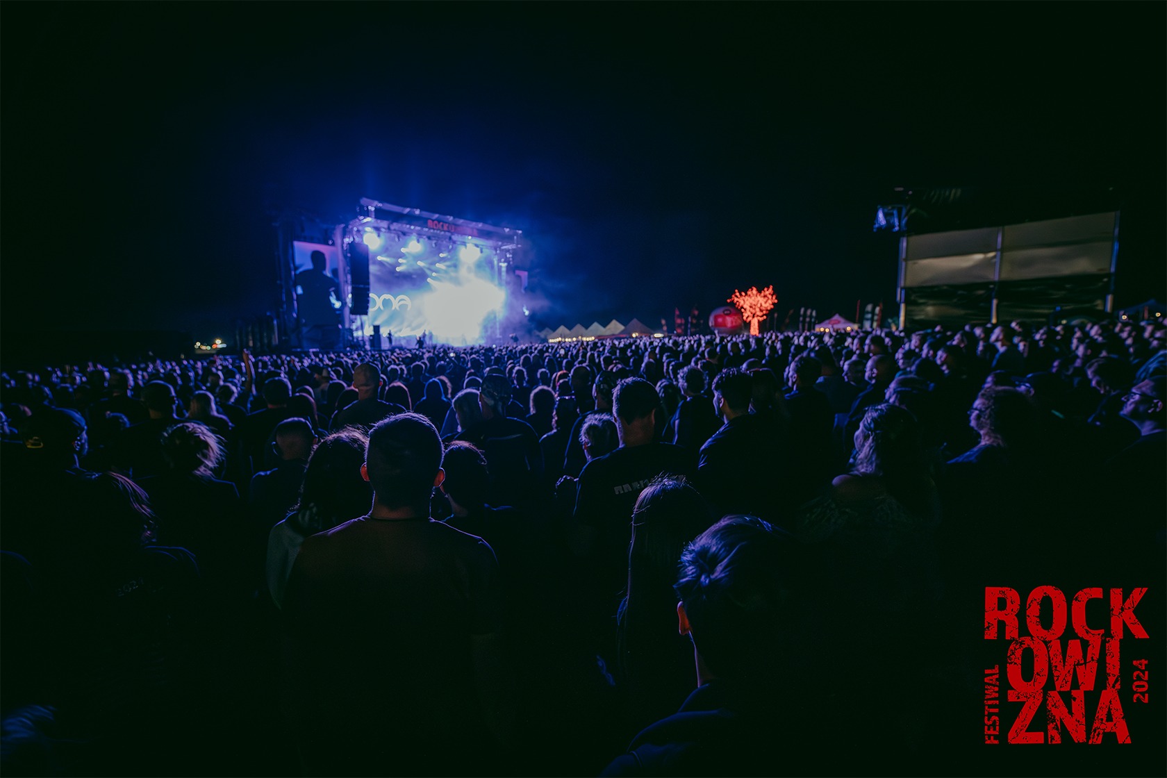 Zdjęcie przedstawia tłum ludzi na koncercie, stojących przed sceną. Zdjęcie zrobiono w nocy.