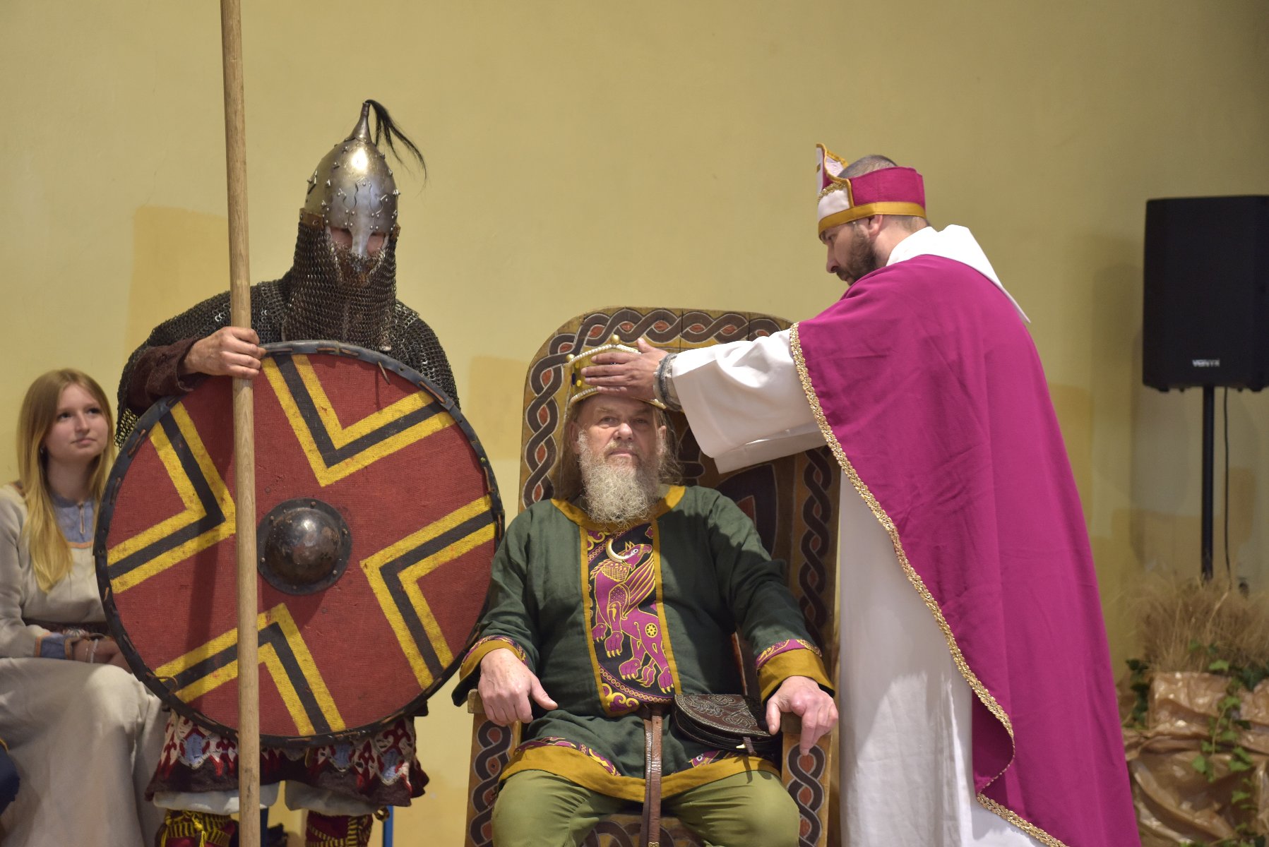 Zdjęcie przedstawia trzech mężczyzn z przebraniach średniowiecznych. Król siedzi na tronie, biskup zakłada mu koronę, obok stoi wojownik.