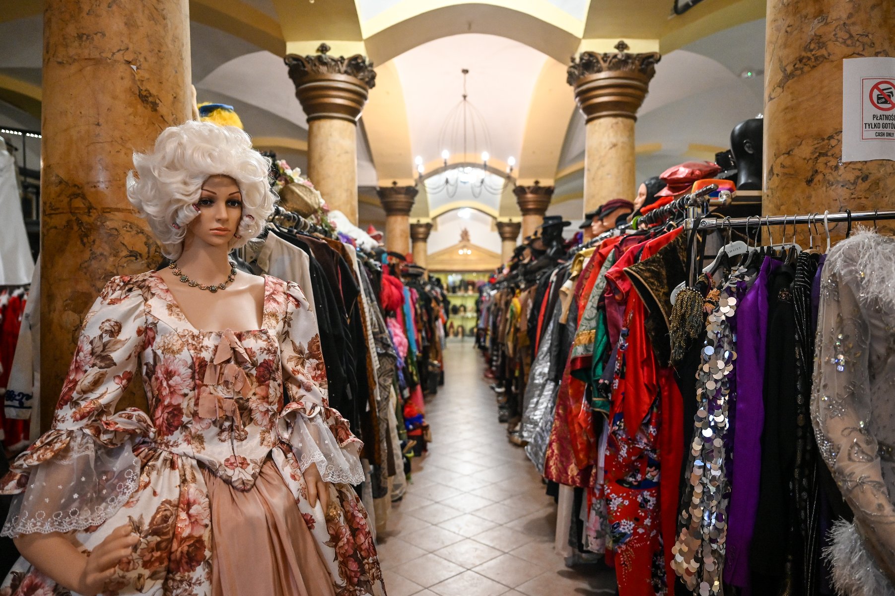 Zdjęcie przedstawia ubrania wiszące na wieszakach w dwóch rządach oraz peruki na półkach.