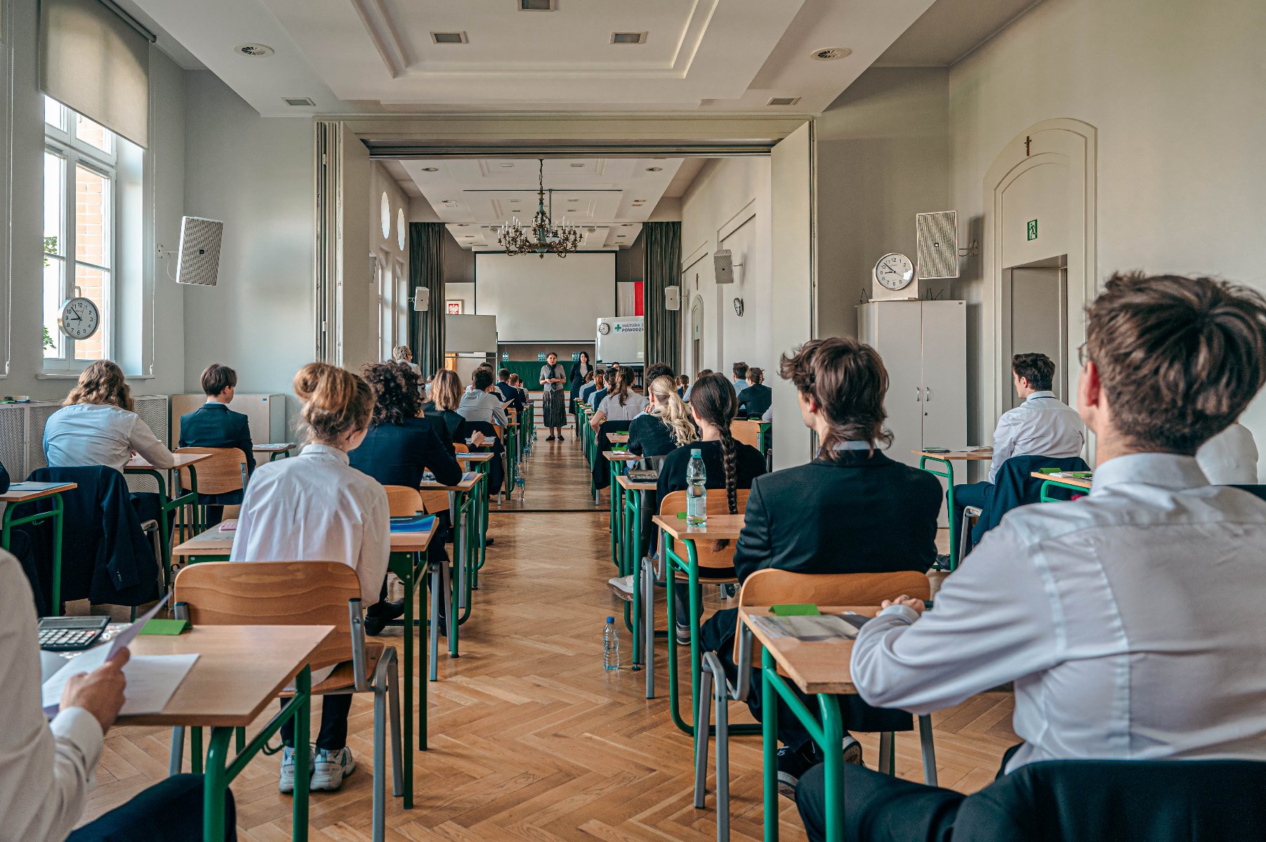 Zdjęcie przedstawia uczniów siedzących w ławkach w szkolnej auli. Młodzież siedzi tyłem do aparatu. Przodem do aparatu i uczniów stoi nauczycielka.