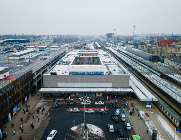 Zdjęcie przedstawia widok z lotu ptaka na stary budynek dworca kolejowego Poznań Główny w zimowej scenerii. Na dachu obiektu, przykrytym cienką warstwą śniegu, widnieje duży niebieski napis z nazwą stacji, a przed wejściem znajduje się półkolisty podjazd, na którym parkują liczne taksówki.