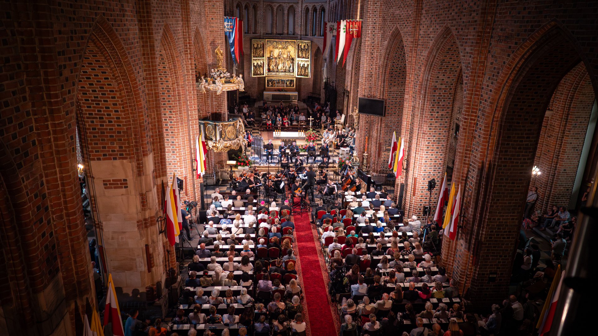 Zdjęcie przedstawia wnętrze katedry. Widać na nim ludzi siedzących w ławkach, a w miejscu ołtarza muzyków z instrumentami. Zdjęcie zrobiono z góry.