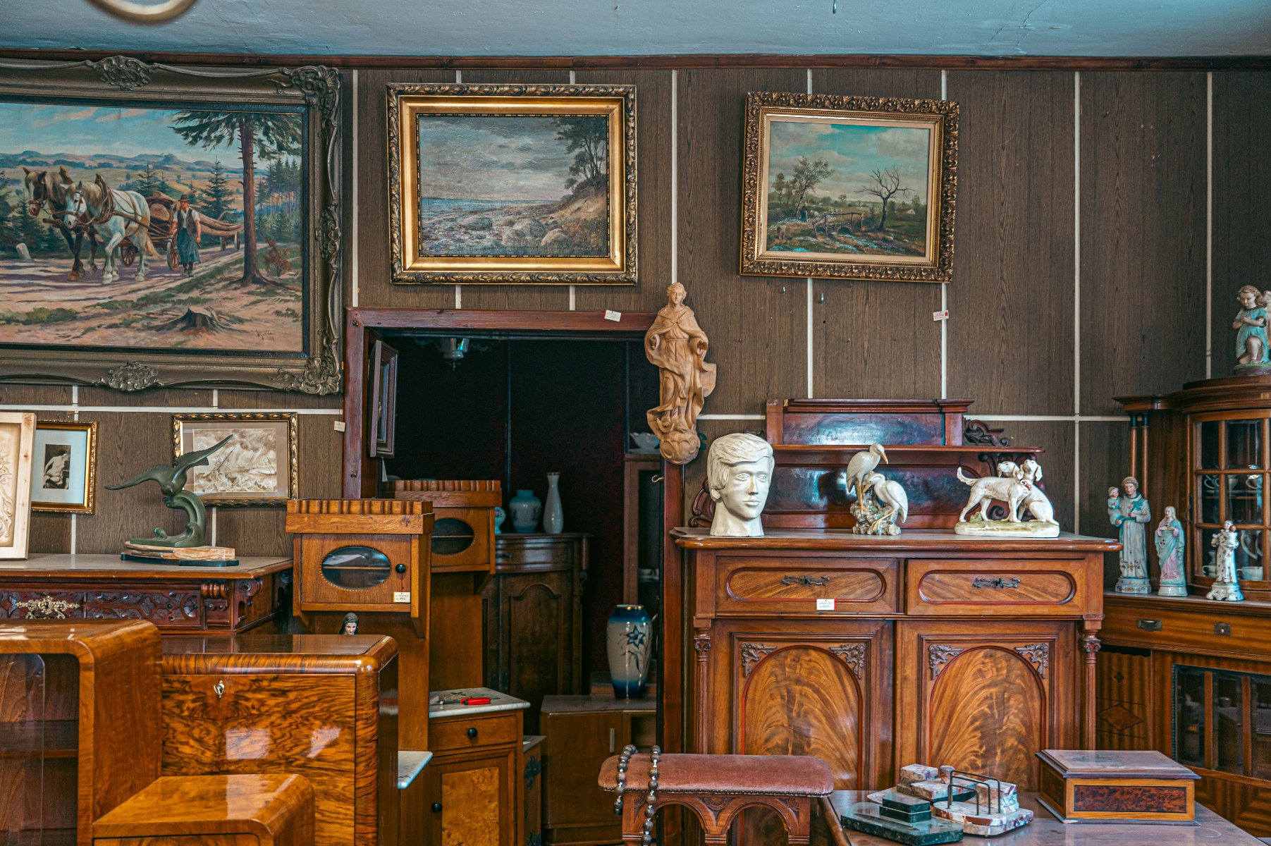 Zdjęcie przedstawia wnętrze sklepu lub pracowni z antykami i przedmiotami kolekcjonerskimi. Pomieszczenie jest gęsto wypełnione meblami, obrazami i różnorodnymi dekoracjami.