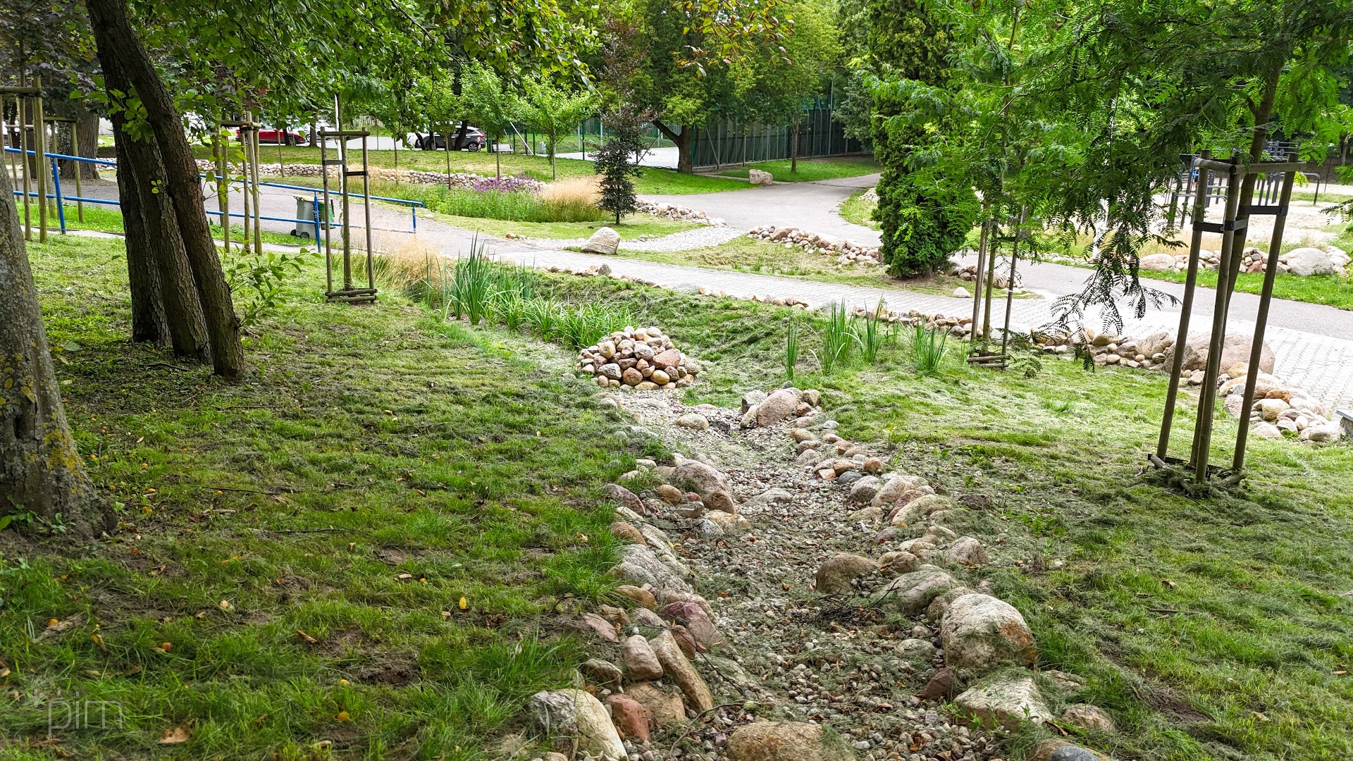 Zdjęcie przedstawia zieleń oraz nieckę wyżłobioną w ziemi. Dalej widać alejki spacerowe.