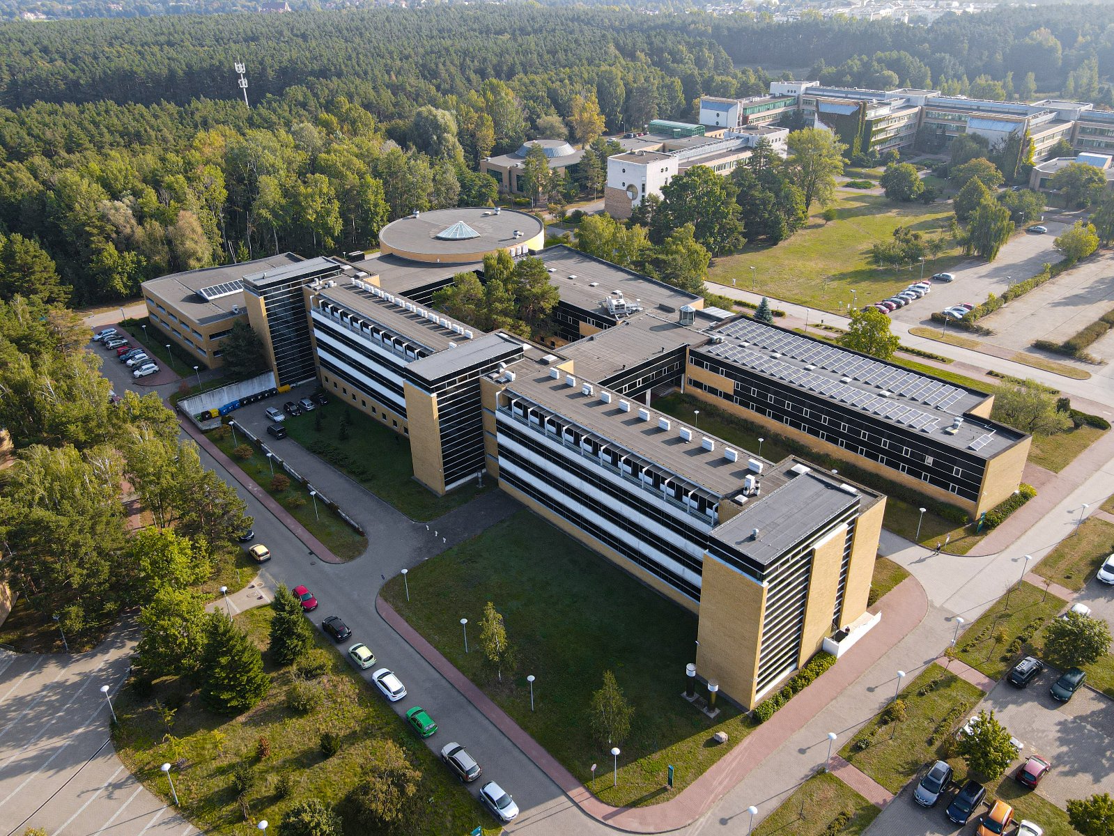 Zdjęcie z drona przedstawiające duży, nowoczesny budynek uniwersytecki z brązową elewacją. Budynek otoczony jest parkingami, ścieżkami oraz zielonymi terenami z drzewami. W tle widoczny jest rozległy las.