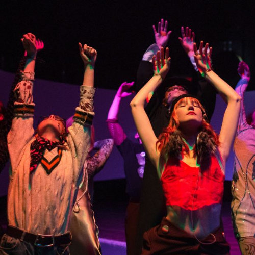 Kilka osób podczas występu. Na pierwszym planie dwie osoby, z podniesionymi rękoma i uniesionymi głowami. Ciemne tło.