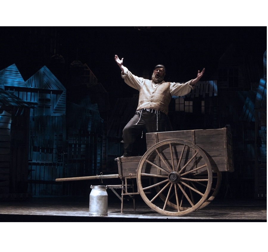 Mężczyzna ubrany w jasną szeroką koszulę, kamizelkę i czarne spodnie stoi na drewnianym wozie. Śpiewa wyciągając ręce przed siebie. Przed wozem stoi kanka na mleko.