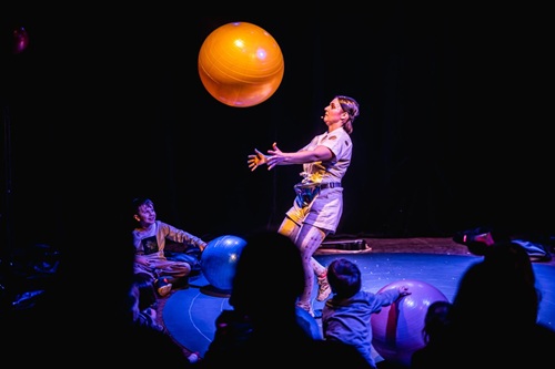 Kobieta na scenie podrzuca dużą pomarańczową piłkę. Widocznych jest kilka obserwujących ją osób. Ciemne tło.