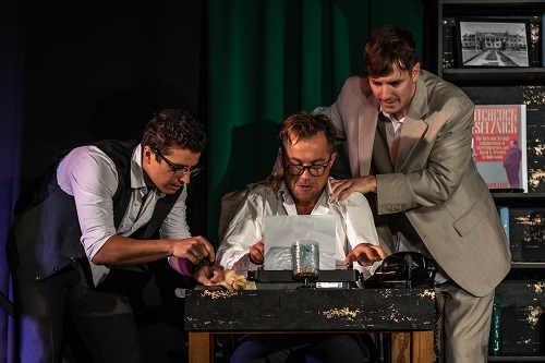 Trzech mężczyzn, jeden z nich siedzi przy stole i pisze coś na maszynie do pisania, Dwóch pozostałych stoi obok niego - jeden z nich trzyma ręce na barkach piszącego.