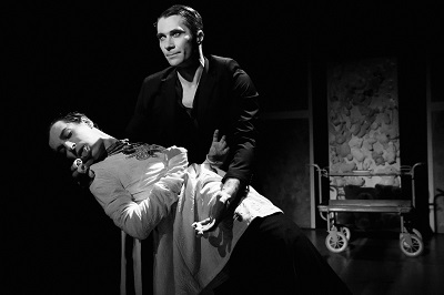 Czarno-białe zdjęcie, na którym jeden mężczyzna podtrzymuje drugiego mężczyznę. Ciemne tło, na drugim planie czterokołowy wózek kelnerski.