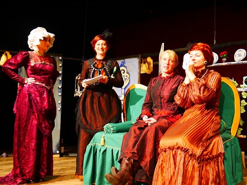 Scena ze spektaklu: cztery kobiety w strojach długich sukniach, dwie z nich siedzą na zielonej sofie, dwie stoją.