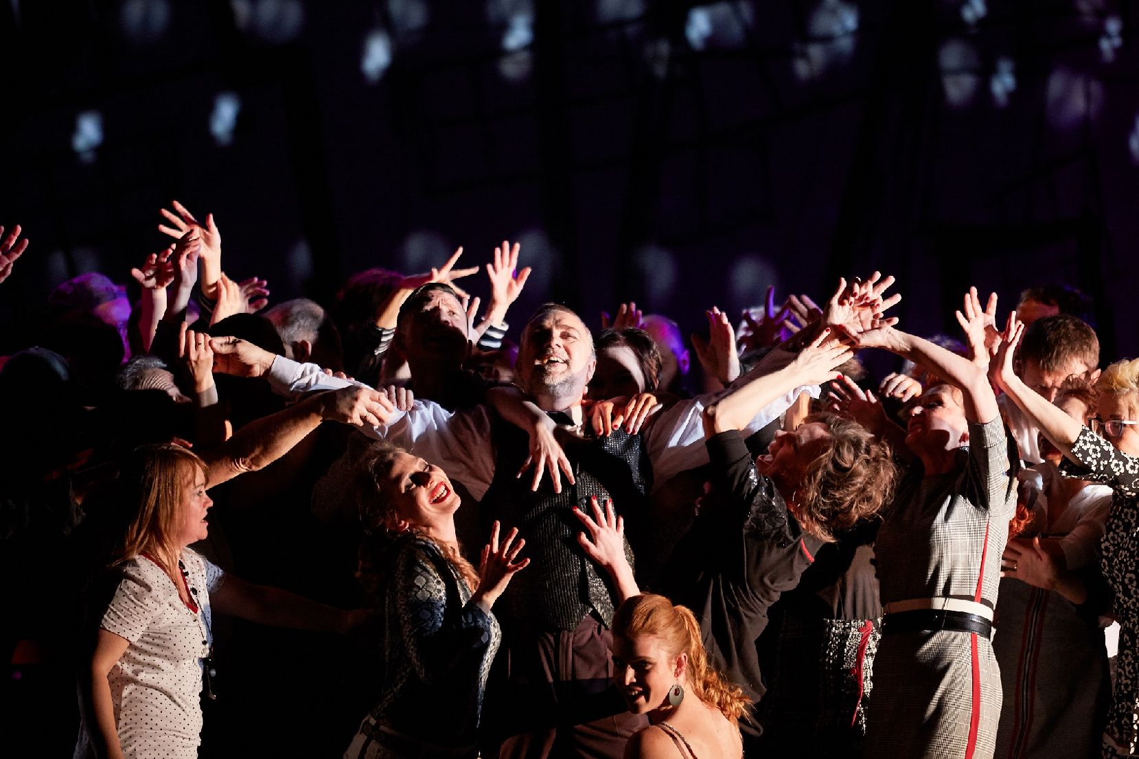 Pośrodku sceny stoi mężczyzna, wznosi ręce w górę, śmieje się, ciasno otacza go tłum ludzi, którzy starają się go dotknąć, śmieją się