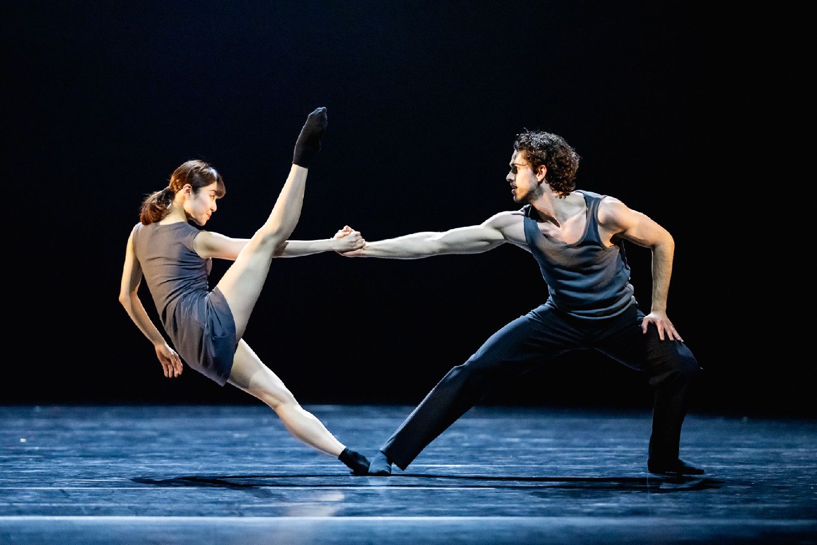 Na scenie kobiet ai mężczyzna w trakcie tańca baletowego. Kobieta stoi naprzeciwko mężczyzny, trzyma go za rękę, jedną nogę podnosi wysoko do góry. Mężczyna trzyma ją mocno, pochylając się do przodu..