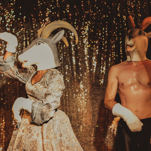 Dwie postacie na scenie: mężczyzna z nagim torsem i kobieta w sukni w srebrno-złotych kolorach. Aktorzy mają założone głowy kozłów. W tle mieniąca się tkanina.
