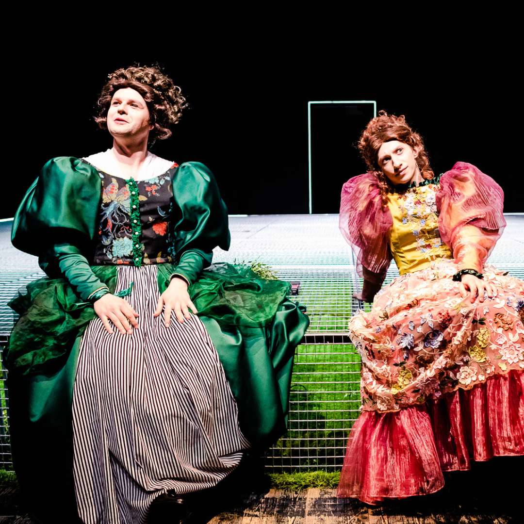 Dwie kobiety w długich, kolorowych sukniach, z bufiastymi rękawami siedzą ma scenie. W tle czarna ściana z białym zarysem drzwi.