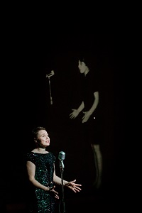 Kobieta w czarnej sukience stoi przed mikrofonem, za nią zdjęcie innej kobiety stojącej przed mikrofonem na czarnym tle.