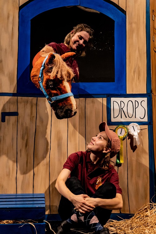 Sena z przedstawienia: przy drewnianej ścianie siedzi kobieta, która patrzy na konia (pacynkę) wystawiajacego łeb przez otwór w ścianie.