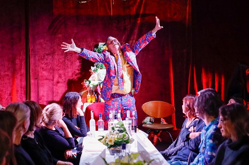 Przy stole siedzi kilka osób, u szczytu stołu stoi mężczyzna w kolorowym garniturze z szeroko rozłożonymi rękoma. Na stole kilka butelek alkoholu.