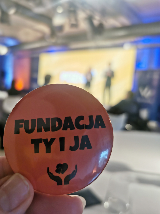 Dłoń trzymająca przypinkę z napisem "Fundacja ty i ja". Tło rozmazane.