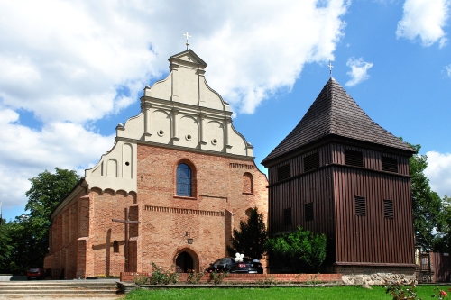 Zabytkowy kościół z cegły obok którego stoi drewniana dzwonnica. Fasada kościoła ma jasny szczyt. Dzwonnica ma ciemne, drewniane ściany. W tle widać niebo z białymi chmurami i drzewa otaczające budynki.