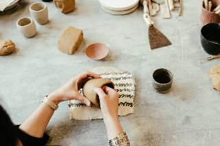 Osoba formuje naczynie z gliny na stole roboczym, na którym leżą różne narzędzia i kawałki gliny.