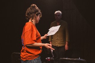 Kobieta oraz mężczyzna w półmroku na scenie. Kobieta jest zwrócona bokiem i w lewej ręce trzyma kartkę. Mężczyzna patrzy na kobietę na wprost.