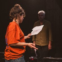 Kobieta oraz mężczyzna w półmroku na scenie. Kobieta jest zwrócona bokiem i w lewej ręce trzyma kartkę. Mężczyzna patrzy na kobietę na wprost.