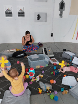 Mamy z dziećmi siedzące na podłodze. Wokół rozrzucone zabawki.