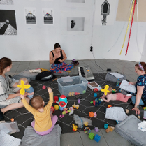 Mamy z dziećmi siedzące na podłodze. Wokół rozrzucone zabawki.