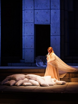 Oświetlona kobieta w długiej, białej szacie na scenie stoi przy położonych na ziemi kilkunastu białych poduszkach. Reszta sceny w kolorach czarno-niebieskich.