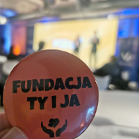 Dłoń trzymająca przypinkę z napisem "Fundacja ty i ja". Tło rozmazane.