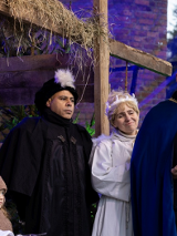 Plenerowa scena jasełek. Widać postacie w historycznych strojach stoją przy drewnianej stajence z sianem. Maria i Józef z dzieckiem w żłóbku, aniołowie oraz trzej królowie, jeden trzyma ozdobną szkatułkę i gestem zwraca się do publiczności.