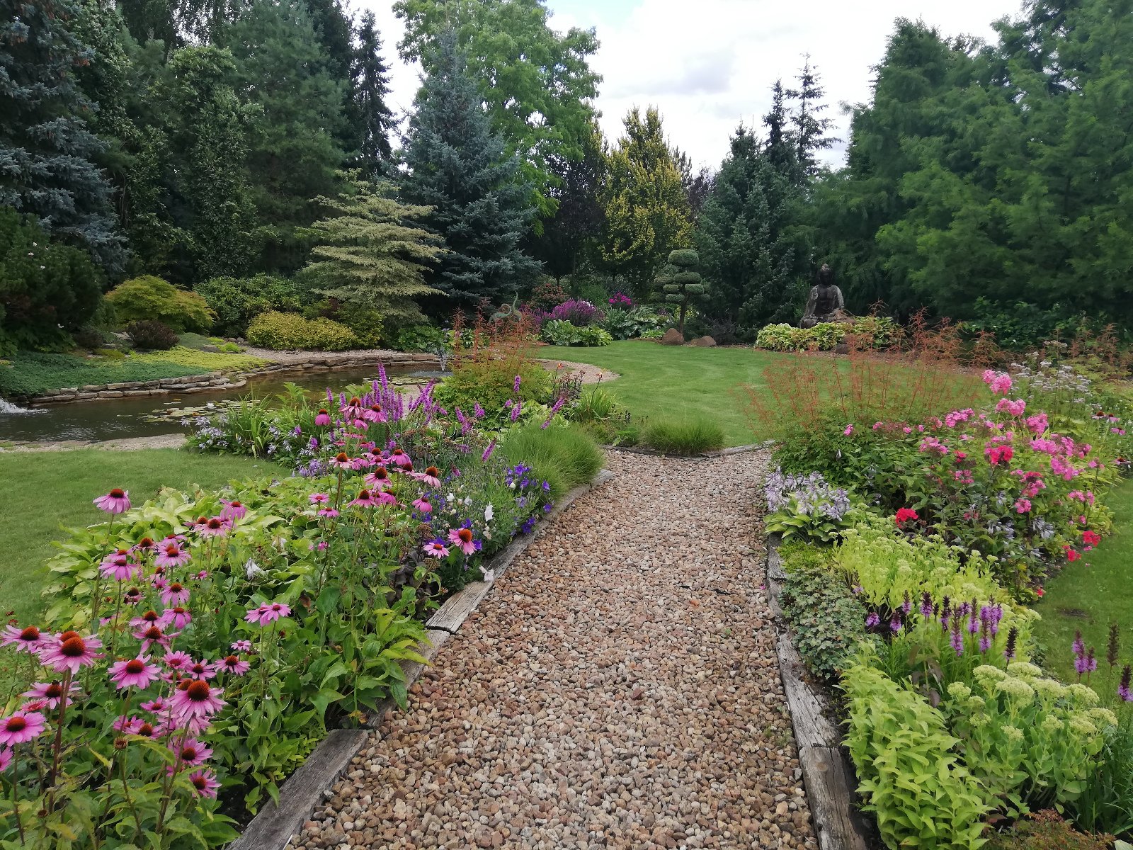 1 miejsce Pani Teresa Dryl-Rydzyńska (Rada Osiedla Krzyżowniki-Smochowice) na pierwszym planie widać ukwieconą alejkę ogrodową z kolorowymi ,różowymi,fioletowymi i czerwonymi kwiatami, królują fioletowe liatry kłosowe, różowe jeżówki,różowe floksy, zielone rozchodniki wyniosłe, w oddali widać oczko wodne z liliami wodnymi i żurawie jako małe ozdoby ogrodowe ,posąg Buddy i rabaty z krzewów ozdobnych i wyższych roślin iglastych.