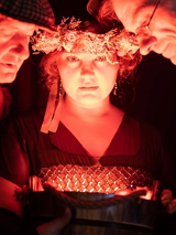 Oświetlone twarze dwóch mężczyzn i kobiety w wianku na głowie. Twarze trojga ludzi są blisko siebie, patrzą na coś w dole.