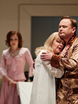 Na pierwszym planie obejmująca się para - kobieta w koszuli nocnej, mężczyzna w szlafroku. W tle kobieta w rózowej sukience, która na nich patrzy.