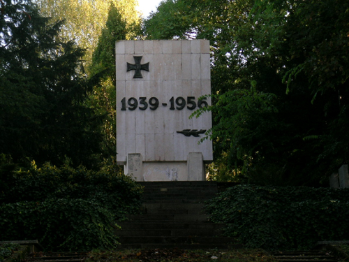 Pamiątkowa tablica z krzyżem Virtuti Militari "1939-1956". Dookoła wysokie drzewa.