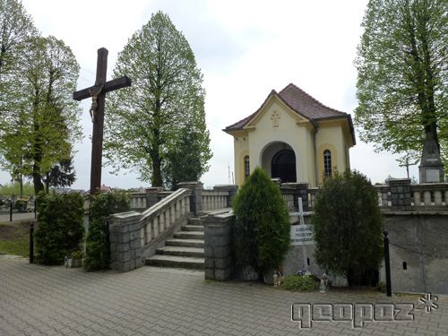 Schody prowadza do kaplicy. Po lewej stronie duży krzyż. Po dwóch stronach kaplicy wysokie drzewa.