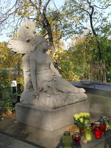 Zabytkowy nagrobek. Rzeźba kobiety, która siedzi i opiera się o krzyż. Na grobie znicze i kwiaty. Dookoła drzewa i inne groby.