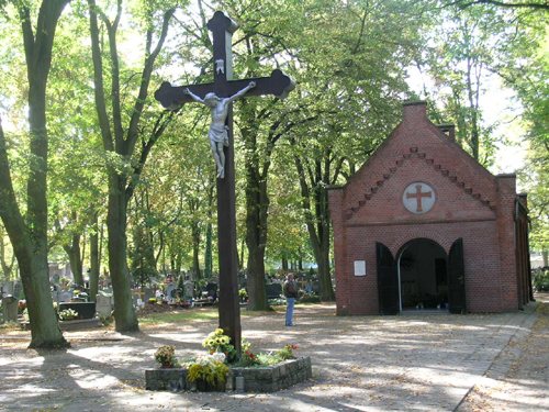 Kaplica na cmentarzu, przed kaplicą nagrobki. Przed kaplicą duży krzyż z figurą Chrystusa. Dookoła drzewa.