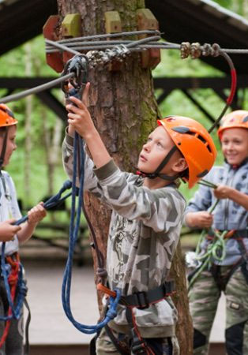 3 chłopców w kaskach w paku linowym.. W głębi stoliki, wiata, drzewa i krzewy.