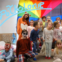 Grupa dzieci z opiekunką robią śmieszne miny. Kilka z nich ma pomalowane twarze. W tle kolorowy materiał.