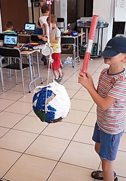 Sala, stoliki i krzesła. Na stołach laptopy. N apierwzym planie chłopiec z akrytymi czapka oczami z piankowa rurka próbuje trafić w zawieszona piłkę. Dalej 3 dzieci przy stolikach.